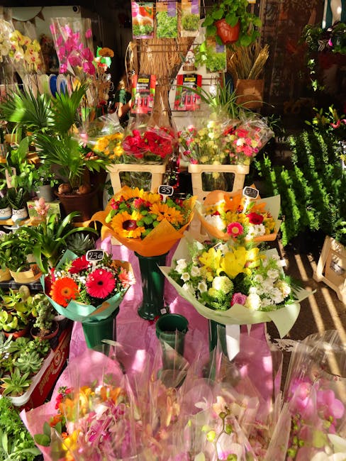 A vibrant display of floral bouquets arranged on a pink cloth-covered surface in a flower shop. The bouquets feature a variety of fresh flowers, including sunflowers, gerbera daisies, and roses, wrapp