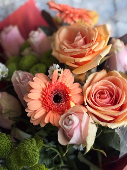 A close-up view of a vibrant flower bouquet featuring peach and pink roses, a coral gerbera daisy, and green chrysanthemums, arranged together with green foliage. The roses have softly ruffled petals,