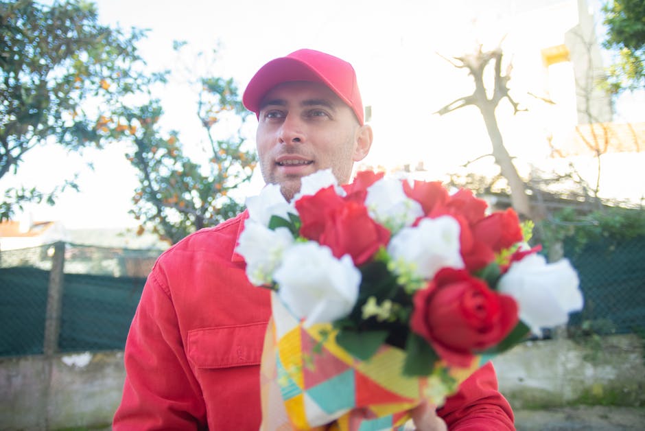 A man dressed in a red jacket and red cap holds a vibrant bouquet of fresh flowers in front of him outdoors, with a blurred background of trees and a fence, illuminated by natural sunlight. The bouque