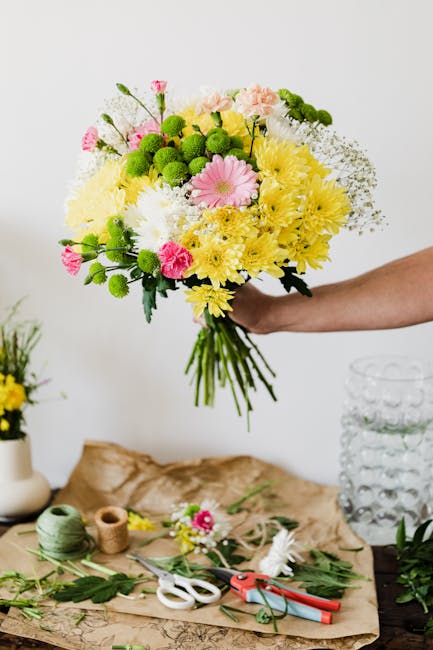 A hand holding a vibrant bouquet of fresh flowers, including pink gerbera daisies, yellow chrysanthemums, white baby's breath, green button mums, and delicate pink carnations, all arranged with lush g
