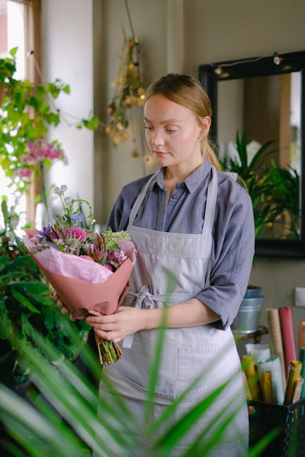 A young woman with light brown hair pulled back, wearing a light grey apron over a blue shirt, is standing indoors in a floristry shop, holding a bouquet of fresh flowers. The bouquet features purple 
