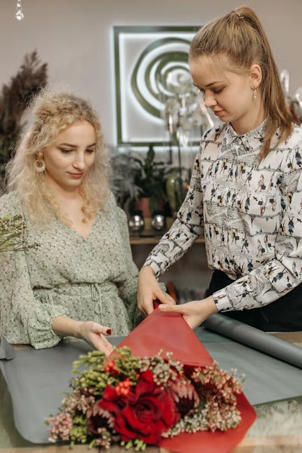 A florist girl with long, light brown hair tied back, wearing a patterned blouse, carefully wraps a bouquet of vibrant red roses, pink and white blossoms, and greenery in red tissue paper. She stands 