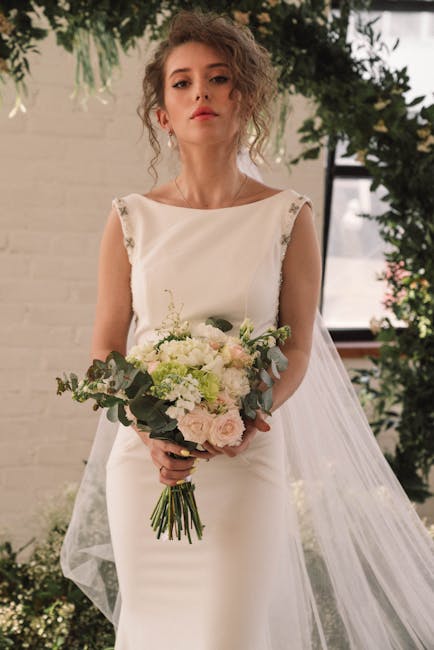 A young woman in a sleeveless, elegant white wedding dress with delicate floral appliqué details on the shoulders, holding a fresh bridal bouquet. The bouquet features soft pastel-colored roses, whit