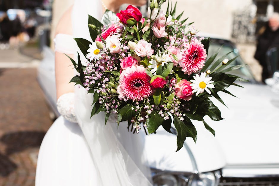 A vibrant bouquet held by a person dressed in white, featuring large pink gerbera daisies, delicate white daisies with yellow centers, and soft pink roses, all surrounded by lush dark green leaves. Th