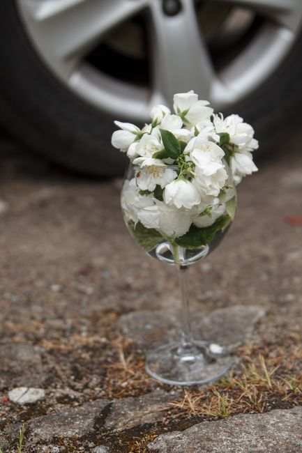 A delicate glass vase filled with fresh white flowers, including lily of the valley and small roses, is placed on a rough, uneven pavement with small stones and patches of dry grass. The bouquet is ar