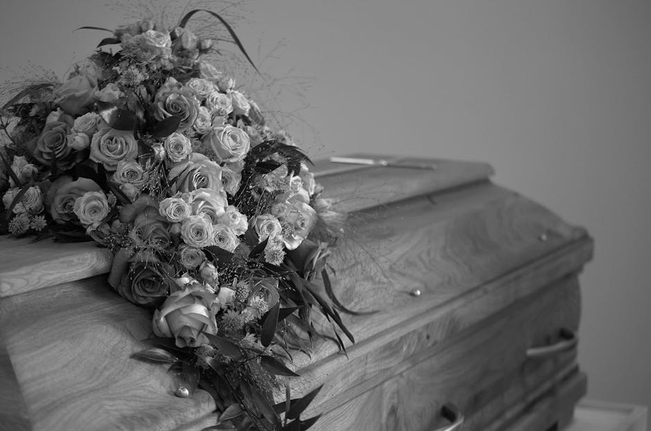 A large funeral bouquet resting on a wooden casket, comprising an arrangement of fresh flowers including roses and other blooms in varying shades of white and cream, with dark greenery and decorative