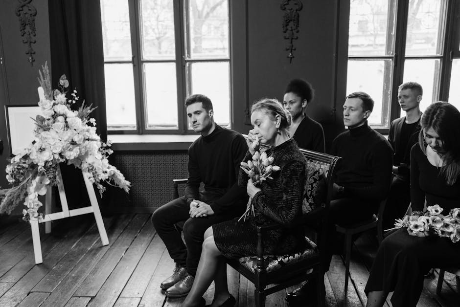 A group of six people seated in a somber indoor setting, attending a funeral or memorial service. Four young men and two women are dressed in dark, formal clothing, with one woman holding a bouquet of