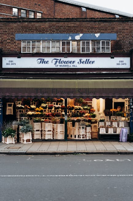 The exterior of 'The Flower Seller' shop on Muswell Hill features a large painted sign and a blue awning above the entrance. The storefront displays an array of fresh floral arrangements and bouquets 